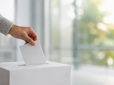 A hand placing a ballot into a ballot box, representing civic participation and voting in local elections.
