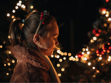 Child standing beside a decorated Christmas tree illuminated with warm white lights during a holiday celebration.