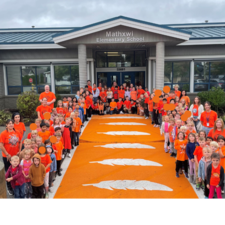 Elementary school students line the orange crosswalk in front of their school wearing orange shirts